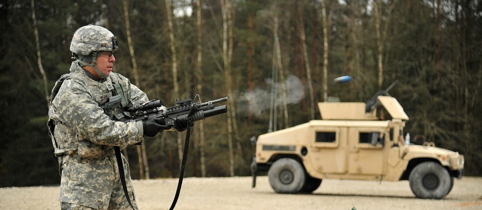 Soldier fires an M203 with a Humvee in the background.