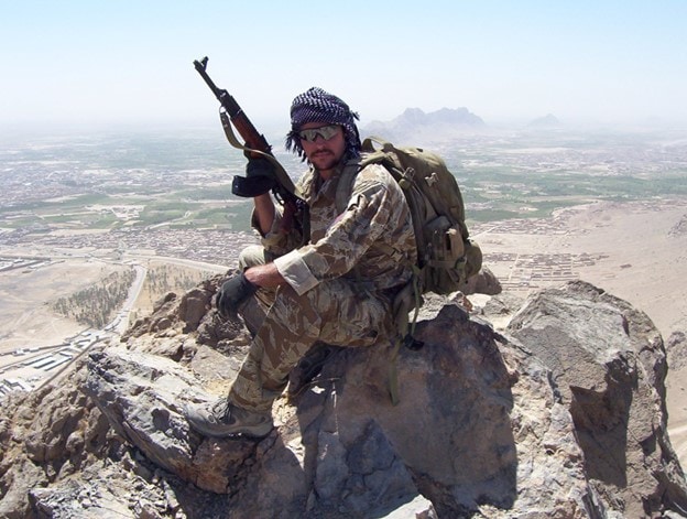 Soldier with an AK sitting on a ridgeline overlooking a desert.