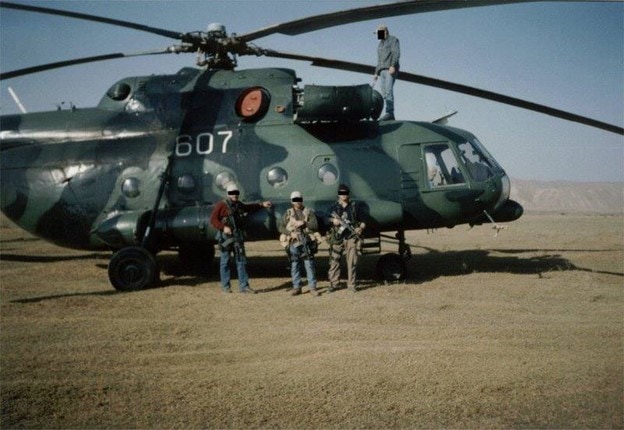 Three soldiers standing infront of a helicopter with a fourth standing on the roof of the cockpit.