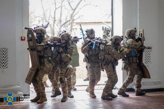 Soldiers enter a building in formation with weapons ready and ballistic shields. 