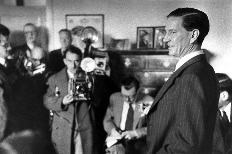 Black and white image of Kim Philby in a room of photographers. 