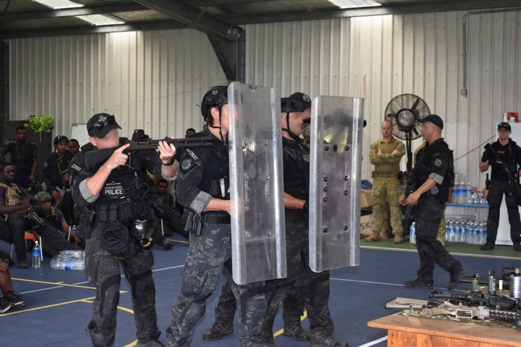 Four Tactical Response Team operators demonstrate training on public order policing tactics using riots shields and shotgun.