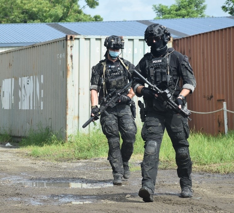 Two Tactical Response Team operators in full kit walking in outdoors police training facility.