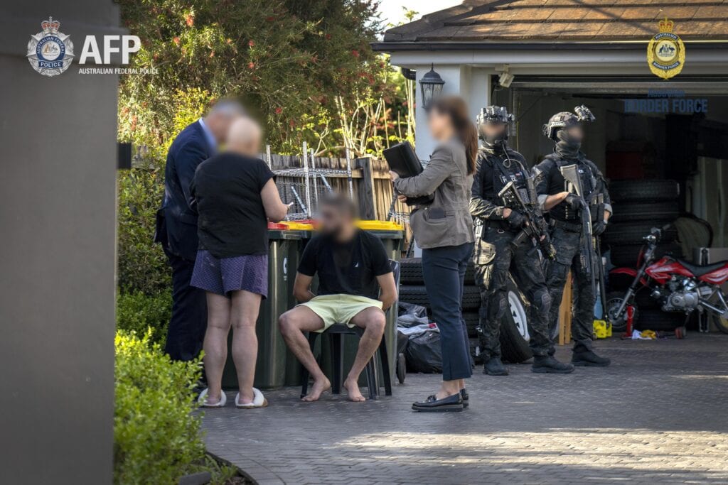 Three AFP police officers and two Tactical Response Team operators stand over arrested suspect outside of home garage.