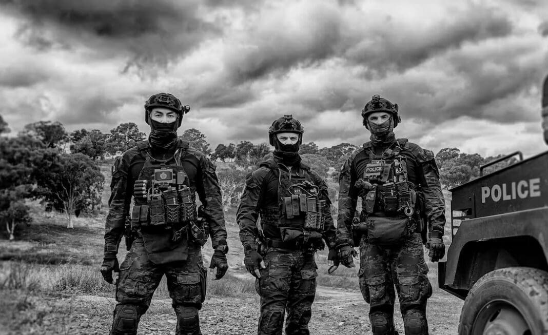 Three Tactical Response Team operators stand in rural environment with police truck to their right.