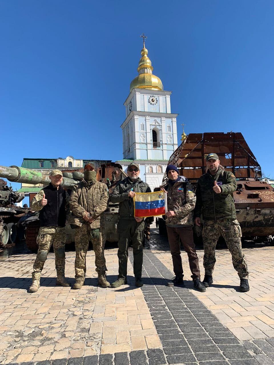 Five men in uniform belonging to the pro-Ukrainian volunteer unit Batallon Bolivar pose with a Venezuelan flag in front of burnt-out Russian armoured vehicles.