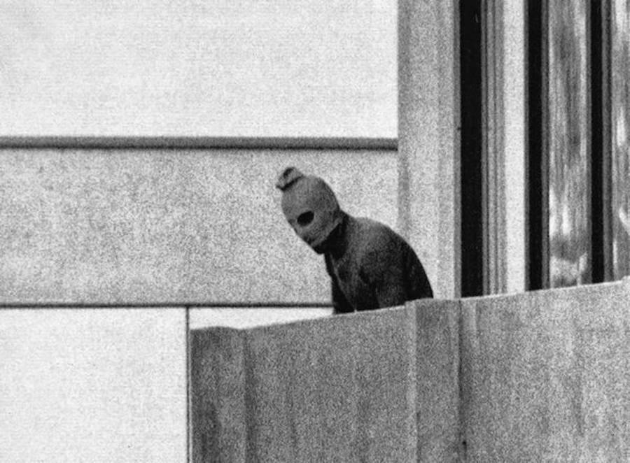 Palestinian terrorist with a balaclava leaning out of the terrace where he stands.