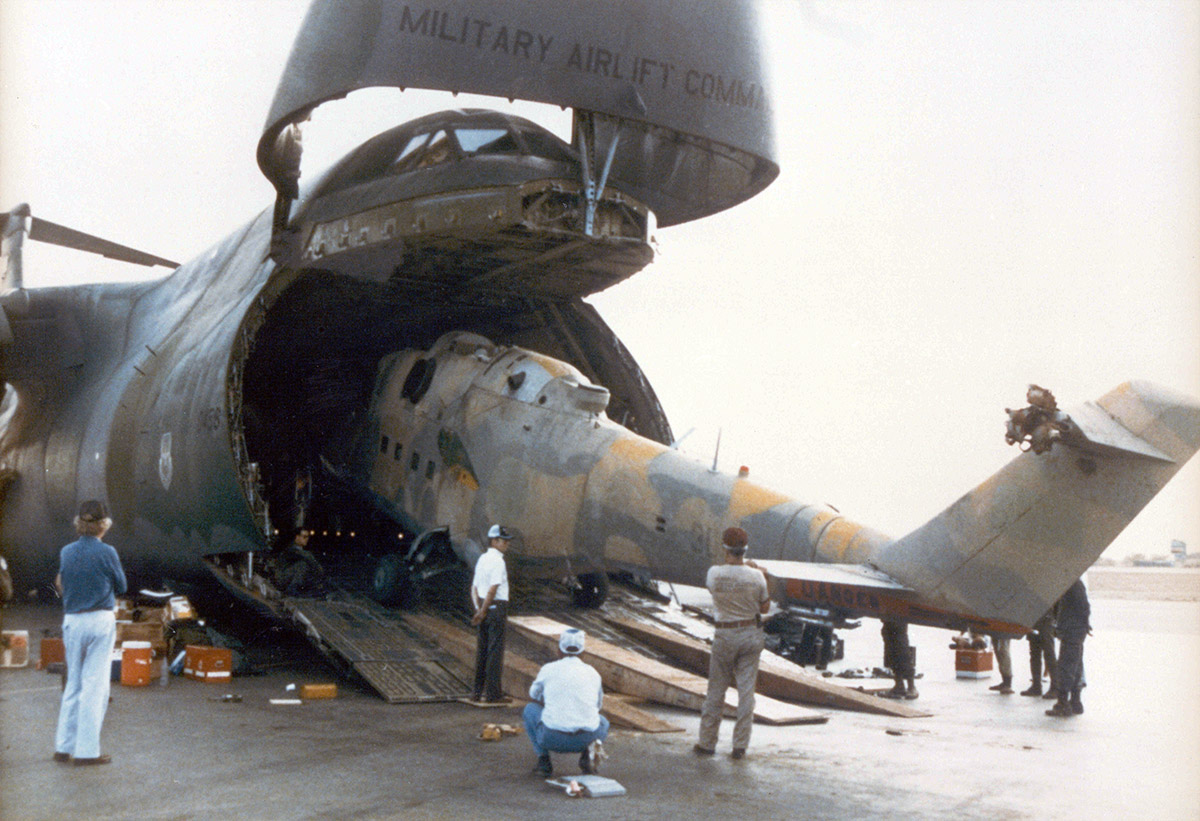 Helicopter being loaded into military cargo plane as people watch.