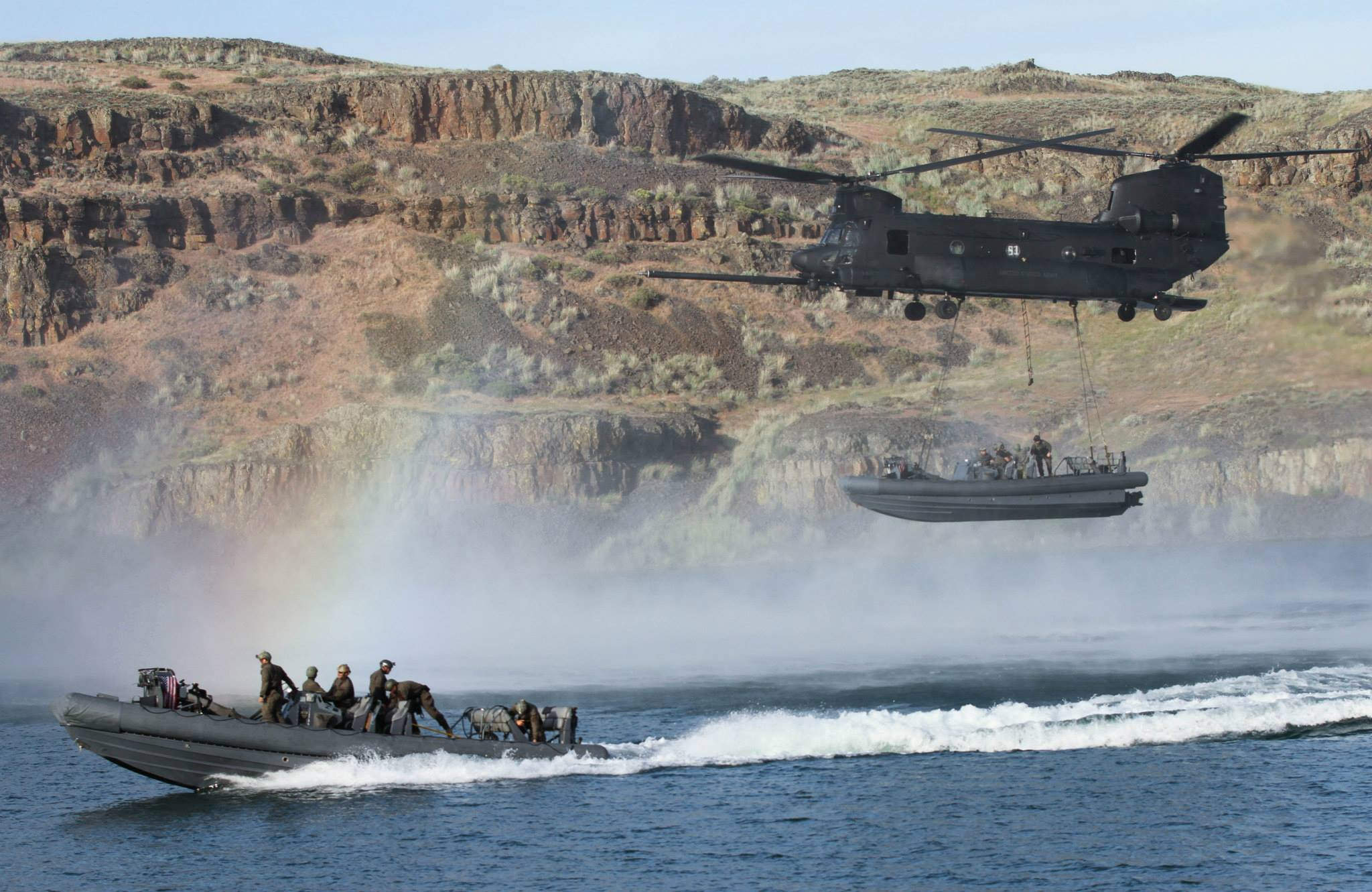 Chinook carrying a boat low above the water via sling load as another boat speeds in the water.